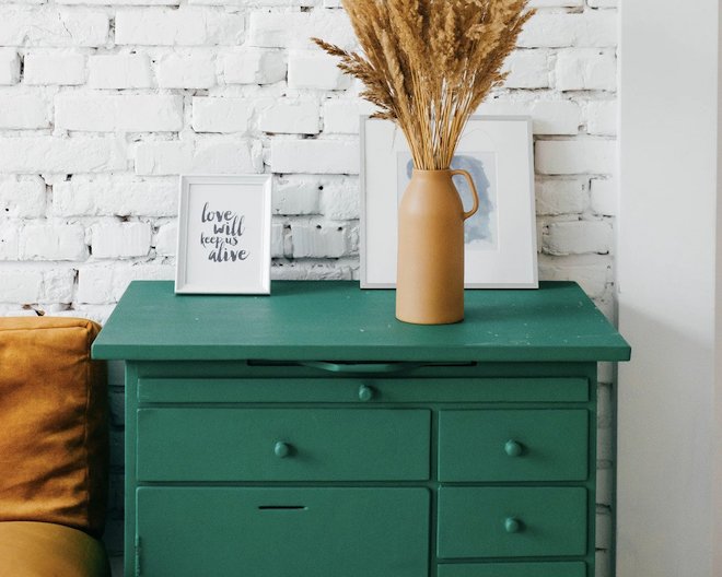 An emerald colored bed side drawer set. On top of the drawer set, lays a painting, a beige colored flower vase, and a frame that says 'Love will keep us alive'